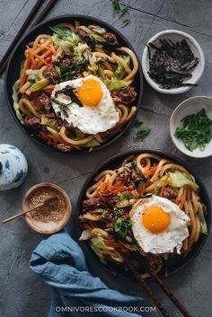 two black bowls filled with noodles and meat topped with fried eggs on top of them