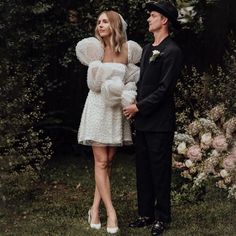 a man and woman standing next to each other in front of some bushes with flowers