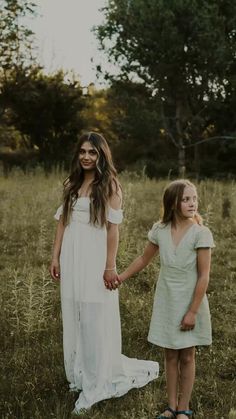 two girls in white dresses holding hands and walking through tall grass with trees in the background