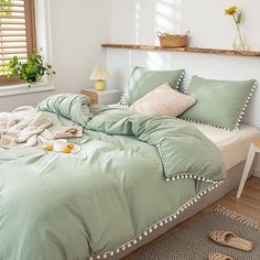 a bed with green comforter and pillows in a white room next to a window