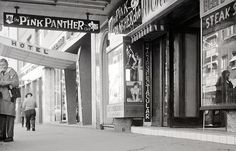 black and white photograph of people walking down the sidewalk in front of shops with signs above them