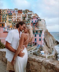 a man and woman standing next to each other in front of the ocean with buildings behind them