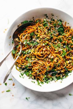 a bowl filled with noodles and vegetables on top of a marble countertop next to a silver spoon