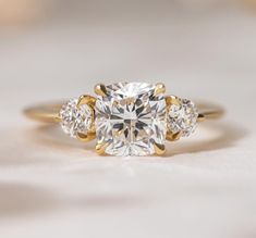 a three stone diamond ring on a white surface with gold trim around the band and sidestones