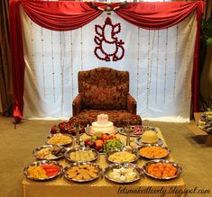 a table topped with lots of food next to a red drapes covered wall and chair