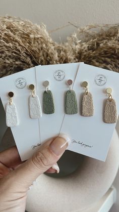 a person holding four pairs of earrings on top of a white table next to a brown teddy bear