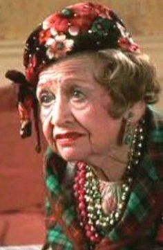 an older woman wearing a red and green plaid shirt with pearls on her head is looking off to the side