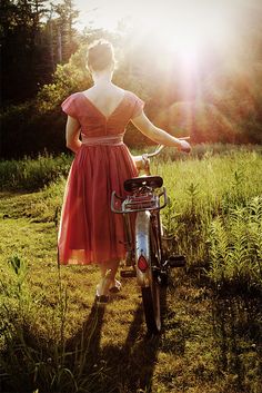 a woman in a red dress standing next to her bike with the sun shining behind her