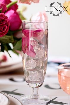 a glass filled with water and ice next to pink flowers on top of a table