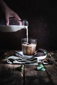 a person pouring milk into a glass filled with ice cream and oreo cookies on a wooden table