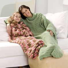 a mother and daughter sitting on a couch wrapped in a crocheted blanket, both smiling at the camera