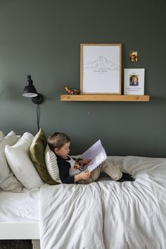 a young boy sitting on top of a bed reading a book next to a lamp