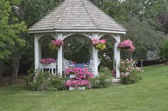 a white gazebo with pink flowers around it in the middle of a grassy area