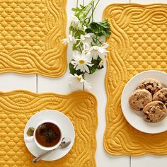 two yellow placemats with white flowers and cookies on them next to a cup of tea