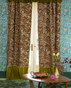 a coffee table in front of a window with green curtains and flowers on the side