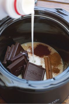 milk being poured into a slow cooker filled with chocolate and cinnamon sticks, along with other ingredients