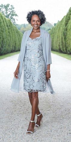 a woman in a blue dress and jacket walking down a path with trees behind her