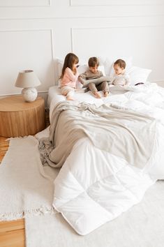 three children are sitting on a bed with white sheets and pillows, reading a book