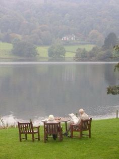 two people sitting at a table on the grass near a body of water with trees in the background