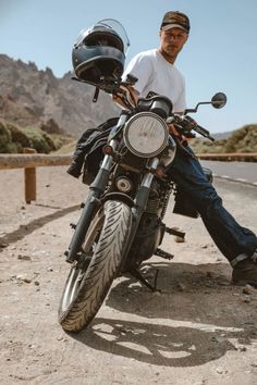 a man sitting on top of a motorcycle in the middle of the desert with mountains behind him