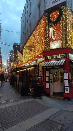 people walking down the street in front of a building with christmas lights all over it