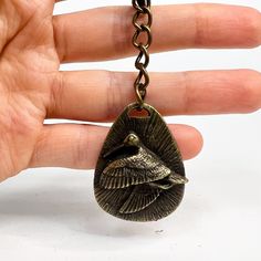 a hand holding a bronze bird shaped keychain on a black chain with a white background