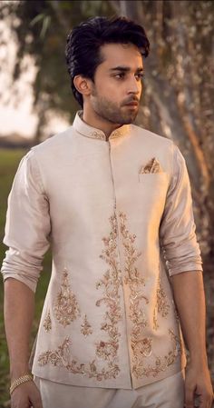 a man standing in front of a tree wearing a white suit with gold embroidery on it