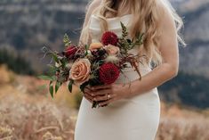 a woman in a white dress holding a bouquet of red and orange flowers with mountains in the background