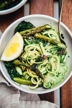 a white bowl filled with zucchini noodles and asparagus next to a lemon wedge