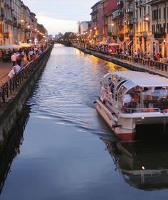 a boat traveling down a river next to tall buildings and people walking on the sidewalk