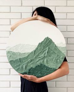 a woman is holding a round painting with mountains in the background and her hands behind her head