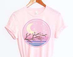 "The Cherry Blossom Sunset Vaporwave T-shirt is perfect for anyone who wants to add a touch of kawaii to their wardrobe. The shirt is made of soft, combed cotton and polyester for a comfortable fit. It's machine-washable and resistant to fading, making it a great choice for everyday wear. The shirt features a beautiful cherry blossom print in shades of pink, purple, and blue. It's perfect for anyone who loves Japanese fashion or lofi vibes. * Unisex * Premium & Soft * Combed & Ringspun Cotton/Po Spring Harajuku T-shirt With Funny Print, Harajuku Style Funny Print T-shirt For Spring, Spring Kawaii Shirt With Graphic Print, Aesthetic Graphic Print Tops For Spring, Spring Aesthetic Graphic Print T-shirt, Spring Aesthetic Graphic T-shirt, Spring Graphic Tee With Moon Print, Spring Moon Print Cotton T-shirt, Vaporwave Aesthetic Outfits