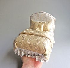 a hand is holding a miniature bed made out of cloths and fabric with gold trim