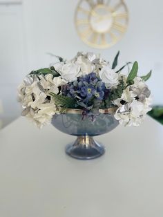 a vase filled with white and blue flowers sitting on top of a table next to a clock