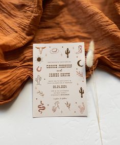 the wedding card is laying on top of an orange cloth covered tablecloth, next to a white feather