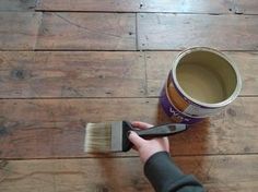 a person is holding a paint brush over a can of brown liquid on a wooden floor