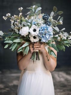 a woman in a white dress holding a bouquet of blue and white flowers with greenery