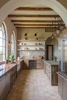 Rustic kitchen with arched windows, wooden beams, tiled backsplash, and open shelves holding pottery. Spanish Villa Kitchen, Spanish Modern Kitchen, Modern Mexican Kitchen, Modern Spanish Decor, Italian Farmhouse Kitchen, Modern Spanish Colonial, Spanish Mediterranean Kitchen, Old Spanish Style Homes, Kitchen Flooring Trends