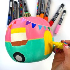 a hand holding a marker next to a painted ball with markers on it and several pens in the background