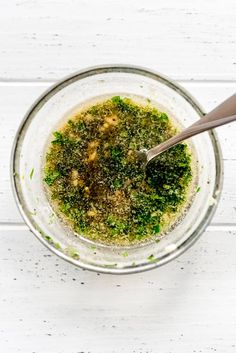 a glass bowl filled with pesto on top of a white wooden table next to a spoon