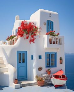 a white house with blue windows and red flowers on the roof, next to a boat