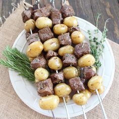 several skewered meat and potatoes on a white plate with rosemary sprigs