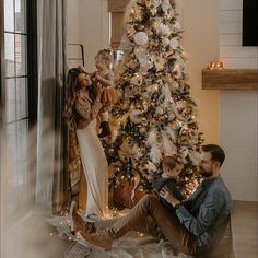 a man sitting next to a woman in front of a christmas tree while holding a baby