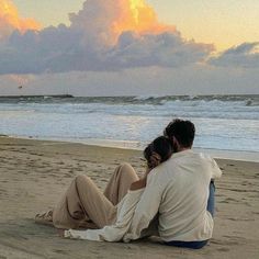 a man and woman sitting on top of a sandy beach next to the ocean at sunset