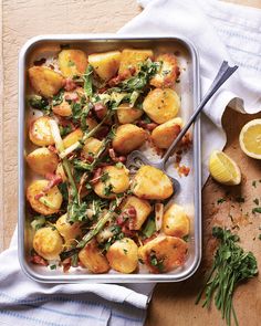 a pan filled with potatoes covered in bacon and green vegetables next to lemon wedges