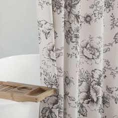 a bath tub sitting next to a shower curtain with flowers on it and a wooden tray