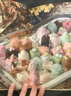 a tray filled with lots of different types of cakes on top of a table next to a person's hand