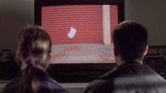 two people sitting in front of a television watching something on the screen with a red brick wall behind them