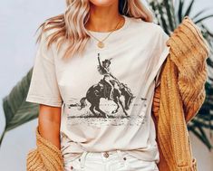 Embrace the spirit of the untamed Wild West with our vintage western cowgirl t-shirt, available exclusively at the Lasso & Lace Etsy shop. This captivating shirt features a stunning vintage cowgirl riding a bucking horse, her hand triumphantly raised in the air against the backdrop of the rugged desert. Captivating Vintage Design: Transport yourself to the rugged landscapes of the Old West with this captivating design. The vintage cowgirl, riding a bucking horse with fearless determination, embo Bohemian Graphic Print Rodeo Top, Bohemian Graphic Print Tops For Rodeo, Relaxed Fit Tops For Country Concerts, Country Style Short Sleeve Tops For Western-themed Events, Vintage Tops For Western-themed Events, Country Style Crew Neck Top For Rodeo, Western Style Graphic Print Shirt For Summer, Casual Graphic Print Shirt For Ranch, Country Style Short Sleeve Top For Concerts