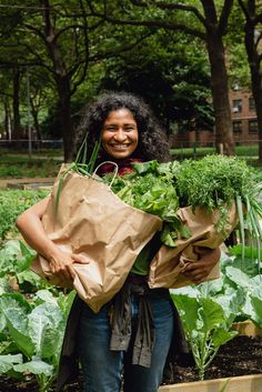 a woman holding bags full of vegetables in a garden with trees and buildings behind her